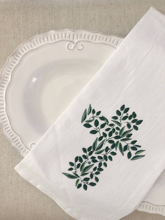 A hand towel featuring a cross out of greenery, folded, and placed on a white plate sitting on a linen tablecloth.